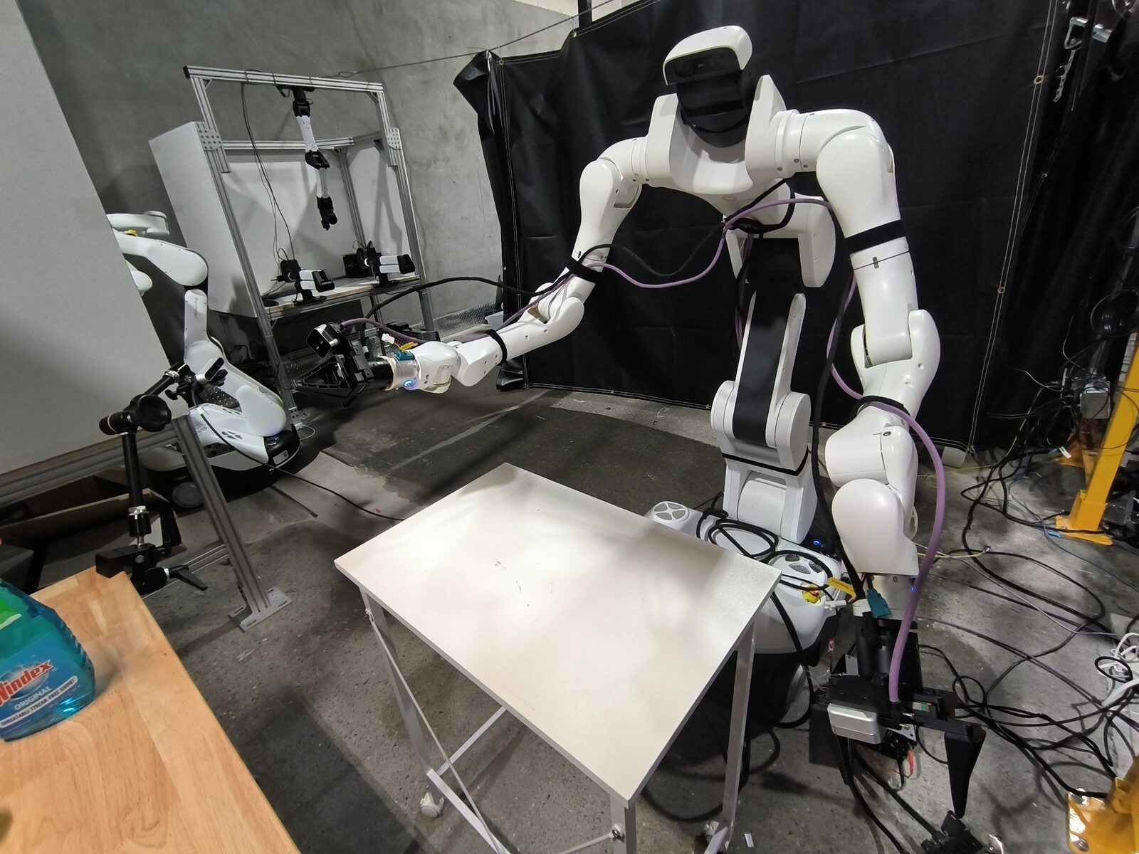 Dexmate Vega Pro wheeled humanoid leaning over a small white table with its wrist camera and gripper extended, a second Vega and camera rig in the background